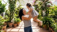 Happy Hispanic mother lifting young daughter in sunny garden patio with lush plants and flowers creating joyful family bonding moment outdoors.