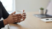 Hands holding white mug at wooden desk with laptop and plant, office break moment for coffee or tea during work day.