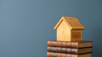 Wooden house model on stack of old books against blue background representing real estate education and property investment knowledge.