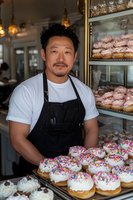 Asian male baker in black apron displaying fresh donuts with colorful sprinkles and frosting in modern bakery. Warm lighting and glass display cases create welcoming atmosphere.