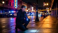 Young Caucasian male police officer crouching on wet city street at night with colorful neon lights and emergency vehicles in background for law enforcement concepts.