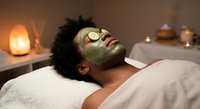 Young African American woman relaxing with green facial mask and cucumber slices on eyes during spa treatment in candlelit wellness center.
