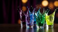Colorful liquid splash in glasses creating dynamic water crown effect with vibrant purple blue green yellow drinks against bokeh background for party concepts.