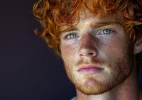 Young Caucasian man with curly red hair, freckles, and green eyes gazes intensely at camera in a close-up portrait on dark background.