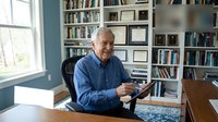 Senior Caucasian businessman working on tablet in home office library with bookshelves and natural window light for remote work concepts.