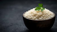 Cooked white basmati rice in dark wooden bowl garnished with fresh parsley on black slate background for healthy cooking and meal preparation.