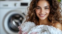 Young mixed-race woman with curly hair smiling while holding fresh white laundry in modern home laundry room with washing machine in background.