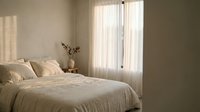 Serene bedroom with cream bedding, neutral walls, and sheer curtains filtering natural light through glass doors, creating a calm, minimalist sleeping space.
