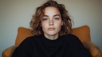Young Caucasian woman with wavy brown hair wearing black turtleneck sweater poses against neutral background. Natural makeup and confident expression create modern portrait.
