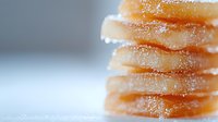 Crystallized ginger slices stacked in soft focus with sugar coating visible on edges, creating delicate layers against blurred light background.