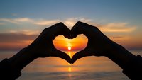 Hands forming heart shape silhouette against golden sunset over calm ocean water. Romantic gesture symbolizing love, connection and peaceful moments by sea.