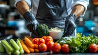 Male chef in black gloves preparing fresh vegetables in professional kitchen. Close-up of hands chopping broccoli with assorted produce including tomatoes, carrots, and celery.