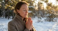 Middle-aged Caucasian woman meditating with prayer hands in snowy winter forest during golden hour sunrise, practicing mindfulness and spiritual wellness outdoors.