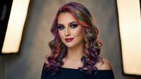 Young woman with vibrant rainbow colored curly hair and bold makeup wearing black off-shoulder top against warm golden lighting background for beauty concepts.