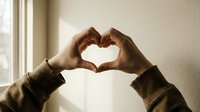 Hands forming a heart shape against a bright window, symbolizing love, care, and connection in warm natural light.