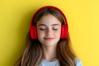 Young woman with closed eyes wearing red wireless headphones enjoying music against vibrant yellow background, expressing serenity and relaxation.