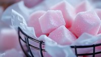 Pink sugar cubes with sparkling crystals in white ceramic bowl with metal grid, macro photography showing sweet texture and delicate details.