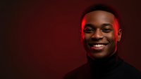 Young African American man smiling confidently in black turtleneck against red background with dramatic lighting for professional headshot photography.