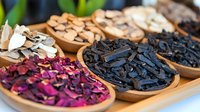 Aromatic dried herbs, flowers and charcoal pieces displayed in wooden bowls on bamboo tray, natural spa and wellness ingredients for aromatherapy and skincare.
