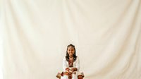 Young woman with long braided hair and gold accessories wearing white shirt, sitting cross-legged against cream fabric backdrop for portrait and lifestyle content.