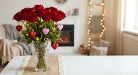 Red roses bouquet in glass vase with heart decorations on white table in cozy living room with fireplace and warm lighting for romantic occasions.