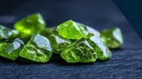 Raw green peridot crystals scattered on dark stone surface with dramatic lighting highlighting natural mineral formation and translucent gemstone texture.
