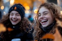 Two young Caucasian women with curly hair laughing outdoors on winter evening, wearing knit hats and scarves, holding coffee cup, with warm bokeh lights in background.