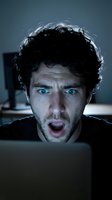 Man with dark curly hair and blue eyes displays shocked expression while working at computer, suitable for business, technology, and emotional reaction concepts.