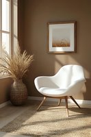 Minimalist interior corner with white modern armchair, dried pampas grass in woven vase, framed wheat field artwork, and natural sunlight casting shadows on brown walls.