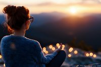 Young woman in cozy sweater watching sunset over mountain landscape with city lights bokeh, rear view silhouette creating peaceful contemplative mood.