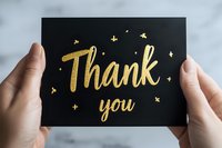 Hands holding elegant black greeting card with golden glittery Thank you text and decorative stars, photographed against blurred light background.