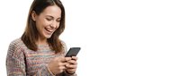 Young woman with dark hair and light skin tone, wearing a pink knit sweater, smiling while using a smartphone. Isolated on white background for web and marketing use.