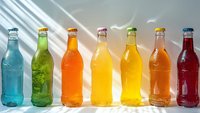 Colorful glass bottles with refreshing drinks arranged in rainbow order with sunlight streaming through, creating a vibrant display perfect for beverage advertising and summer lifestyle content.