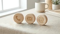 Wooden contact icons with phone, email, and at symbol on desk surface next to coffee mug and plant for business communication and website design.