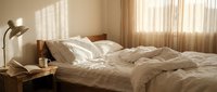 Cozy bedroom interior with unmade bed, warm sunlight streaming through sheer curtains, wooden furniture and soft neutral tones creating peaceful morning atmosphere.
