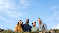 Diverse group of four adults standing outdoors against blue sky with clouds, wearing casual clothing in yellow, navy, green and gray. For lifestyle, community, teamwork and diversity concepts.