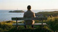 Man sitting on wooden bench overlooking coastal landscape with island and ocean at sunset, contemplative outdoor scene for travel, lifestyle, and wellness content.