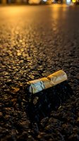Discarded cigarette butt on wet asphalt street at night with golden streetlight reflections creating atmospheric urban scene for anti-smoking campaigns.