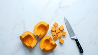 Fresh butternut squash cut in half and diced into cubes with sharp kitchen knife on white marble surface ready for cooking and meal preparation.