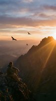 Hiker sitting on mountain cliff at golden hour sunrise watching birds soar over vast valley landscape below with dramatic sunrays piercing clouds.