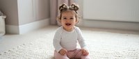 Happy toddler girl with curly hair sitting on floor in bright modern home interior laughing and smiling with joy during playtime.