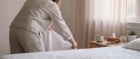 Young woman in beige pajamas sitting on bed edge holding head in hands showing depression, mental health struggles, morning sadness in bright bedroom.