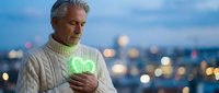 Middle-aged Caucasian man in cream sweater holding glowing green heart symbol at twilight city backdrop with bokeh lights expressing love and connection.