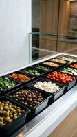 Salad bar display with fresh vegetables, olives, tomatoes, cheese, and greens in black containers behind glass counter in modern restaurant or catering kitchen setting.