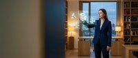 Professional businesswoman in navy suit standing in modern office interior with warm lighting and city view through windows.