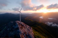 Wind turbine on mountain peak at dramatic sunset with golden light rays illuminating valleys, lakes and rolling hills below, showcasing renewable energy in nature.
