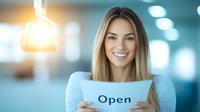 Young Caucasian businesswoman with blonde hair holding open sign in modern office interior with warm lighting and blurred background, expressing welcoming attitude.