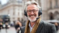 Senior Caucasian man with gray hair and beard wearing headphones with microphone, smiling in urban setting with blurred city architecture background.