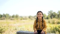 Young girl with braided hair, wearing brown jacket, smiling at camera in rural agricultural field during daytime. Suitable for educational, lifestyle, and community content.