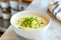 Creamy potato leek soup garnished with fresh chopped chives and melted butter in white ceramic bowl, selective focus on herbs and broth surface.